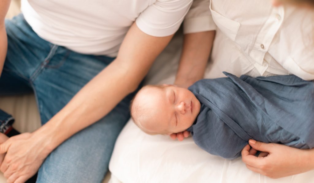 Close-up of parents holding newborn baby son wrapped in swaddle blanket at home.
