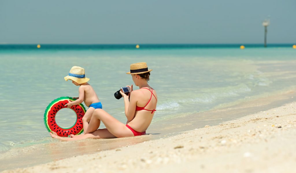 Two year old toddler boy on beach with mother holding camera