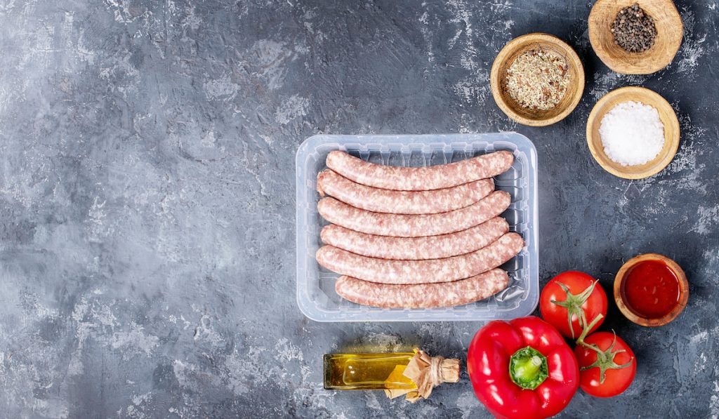 raw pork sausages, bell pepper, tomatoes, oil, and seasoning on dark gray background